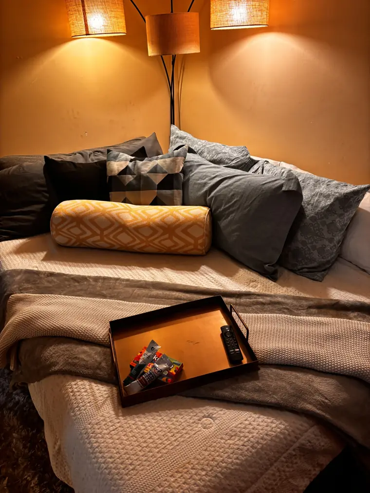 A made bed with layered gray pillows, a yellow bolster, and a serving tray under a three-shade arc lamp